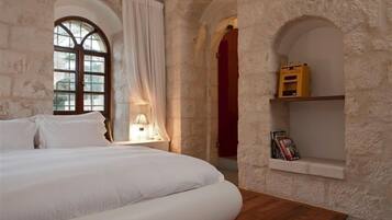 A bedroom with a large bed, a wooden nightstand, a window with curtains, and a stone wall with a shelf.