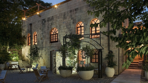 Un edificio de piedra con ventanas arqueadas y una pérgola de madera. Hay zonas de estar al aire libre con sillas y mesas, rodeadas de grandes macetas.