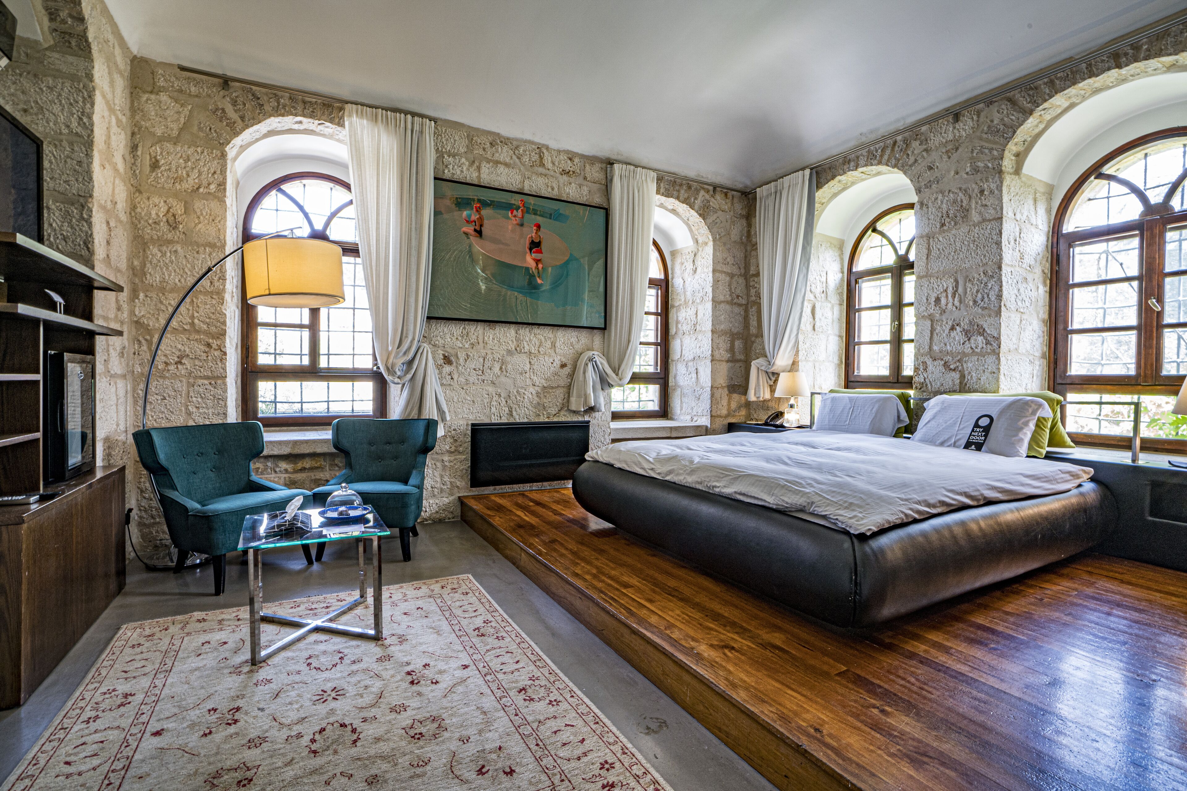 A bedroom with a large bed, two armchairs, a glass coffee table, and a painting on the wall.