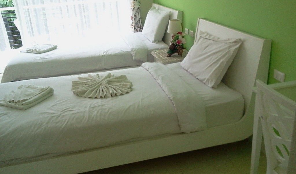 A hotel room with two beds, white bedding, a bedside table with a lamp and flowers, and a green wall.