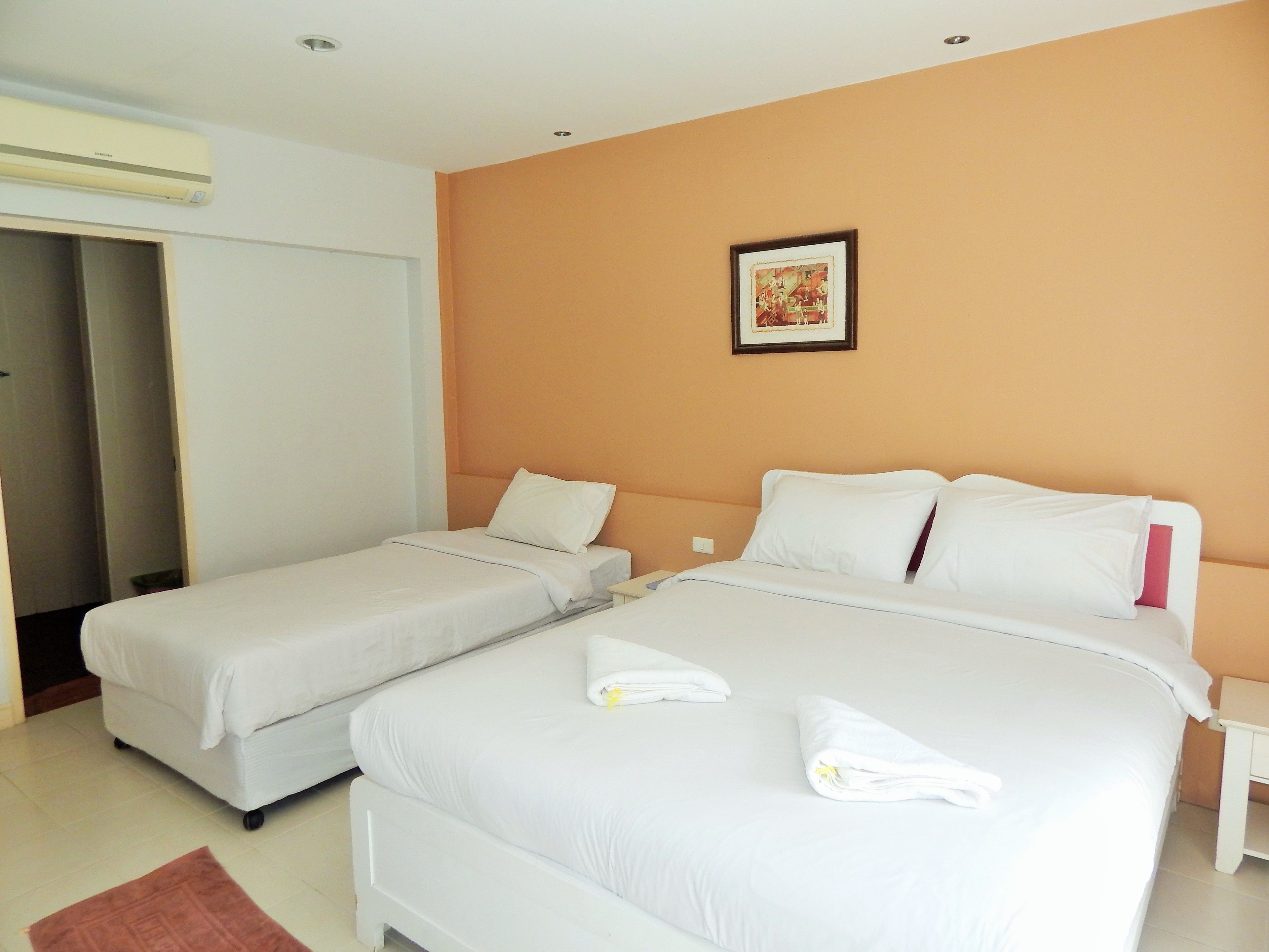 A hotel room with two single beds, a large bed, a white air conditioner, a framed picture on the wall, and a brown rug.