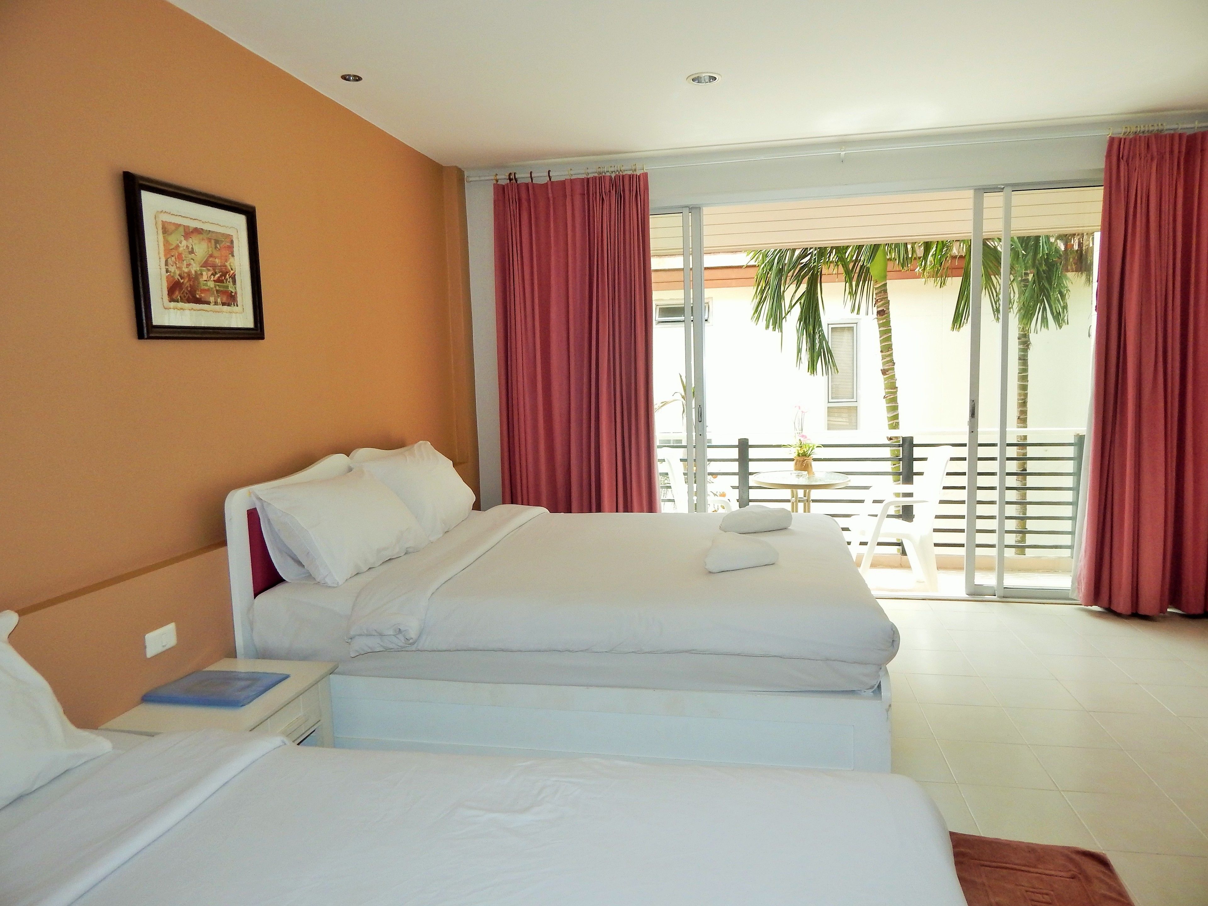 A hotel room with two beds, a balcony with red curtains, and a view of palm trees outside.