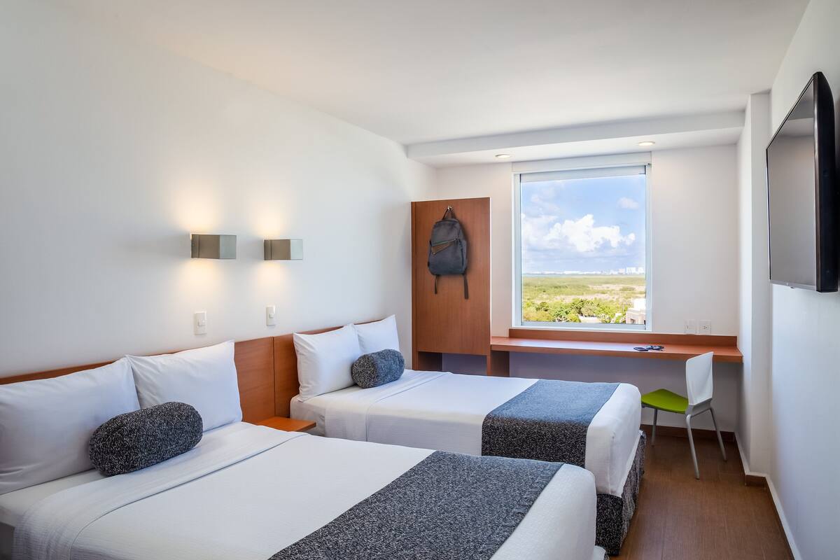 Habitación de hotel con dos camas, un escritorio, una silla y un amplio ventanal con vistas.