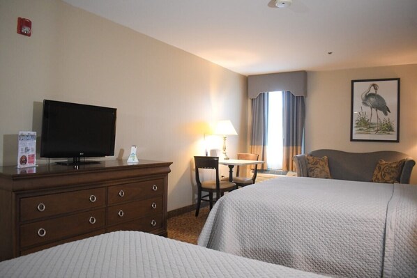 A hotel room with two beds, a television on a dresser, a desk with a lamp, and a window with curtains.