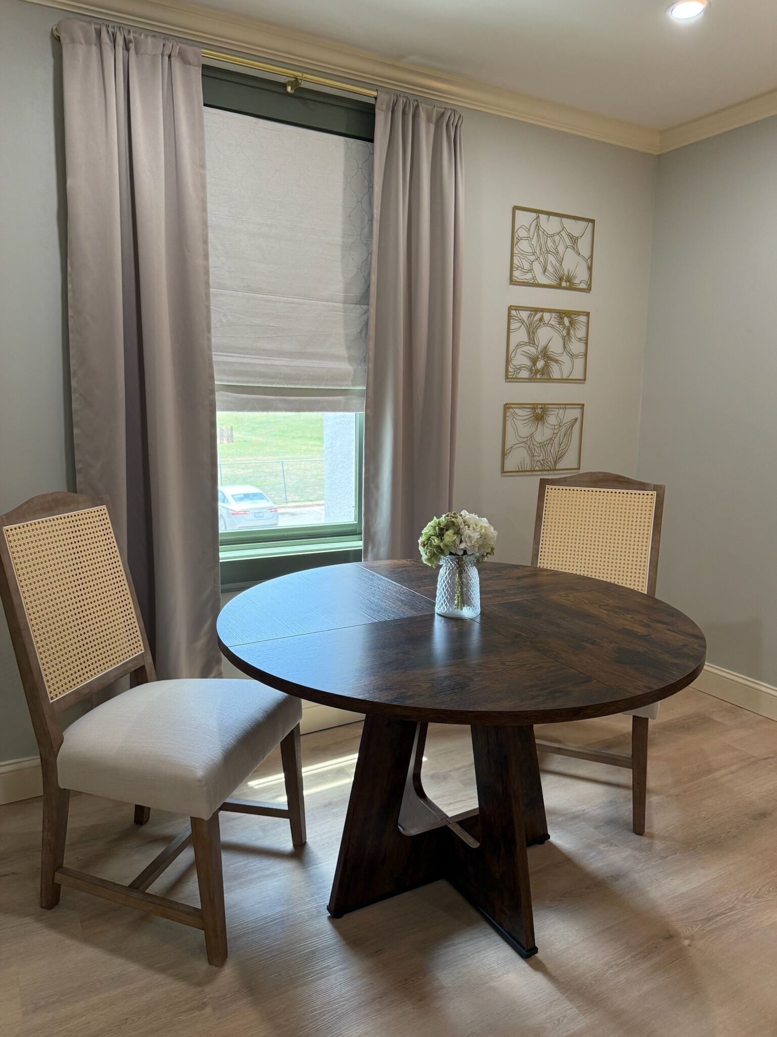 A dining area with a round table, two chairs, a vase with flowers, and a window with curtains.