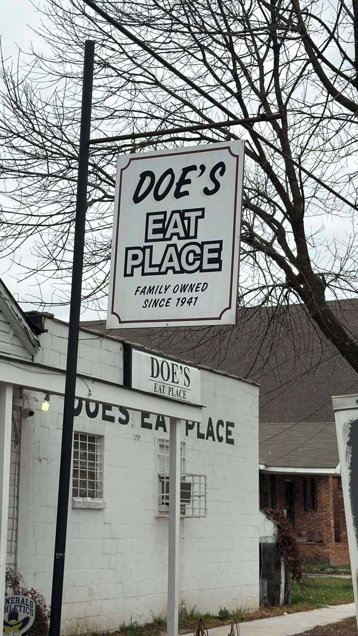 A sign for Doe's Eat Place, a family-owned establishment since 1941, with a white building and bare trees in the background.