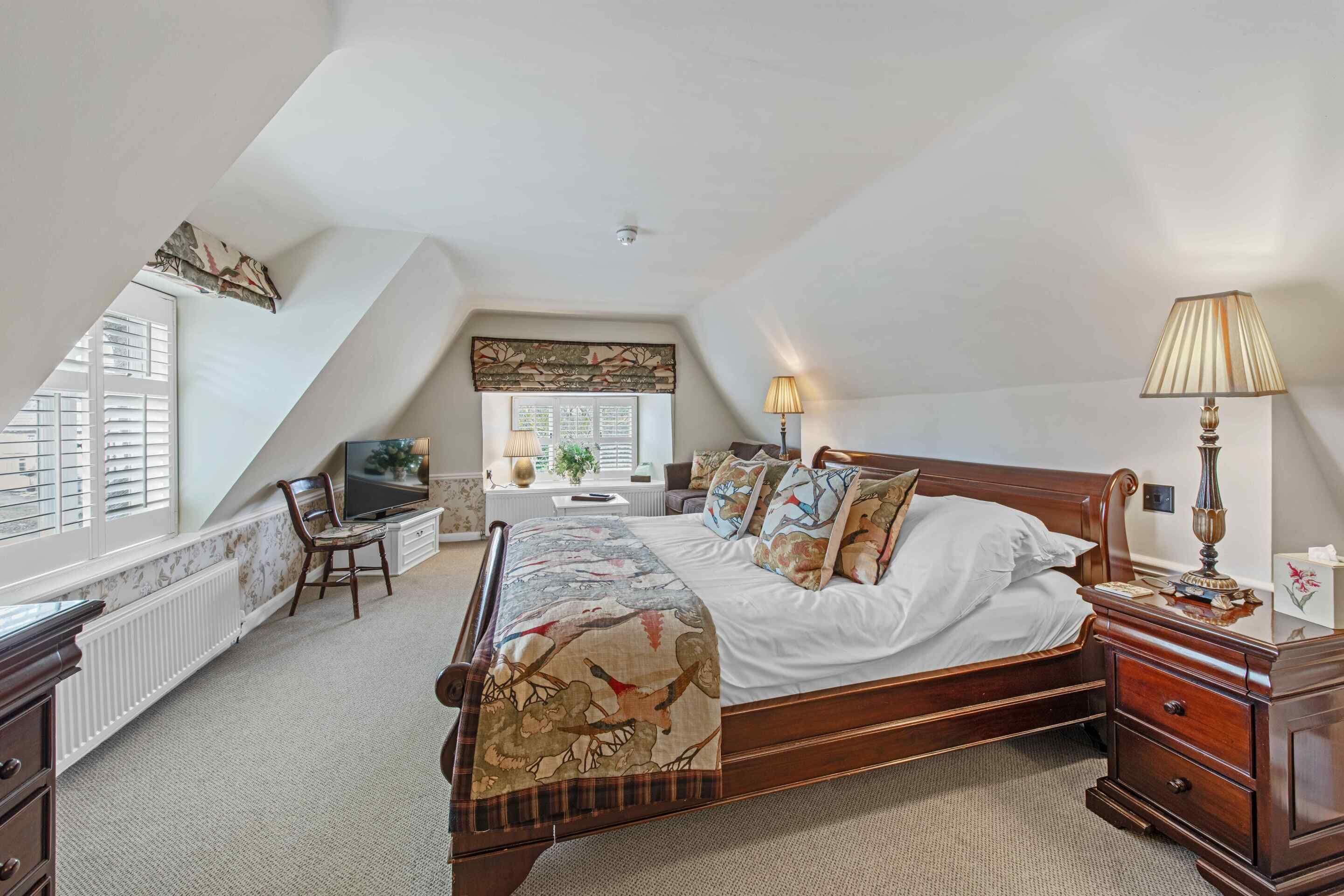 A bedroom with a sloped ceiling, a bed with a patterned bedspread, a bedside table with a lamp, a chair, and a television.