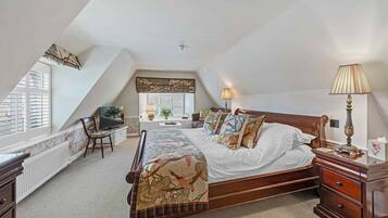 A bedroom with a sloped ceiling, a bed with a patterned bedspread, a bedside table with a lamp, a chair, and a television.