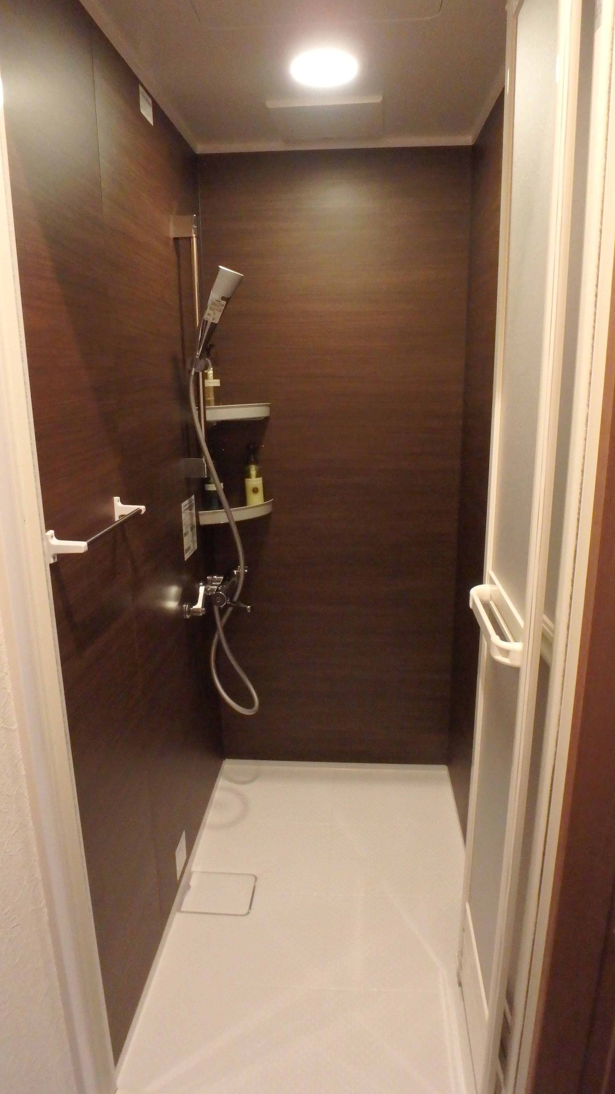 A small, narrow shower room with a wooden door, a white shower stall, and a shelf with toiletries.