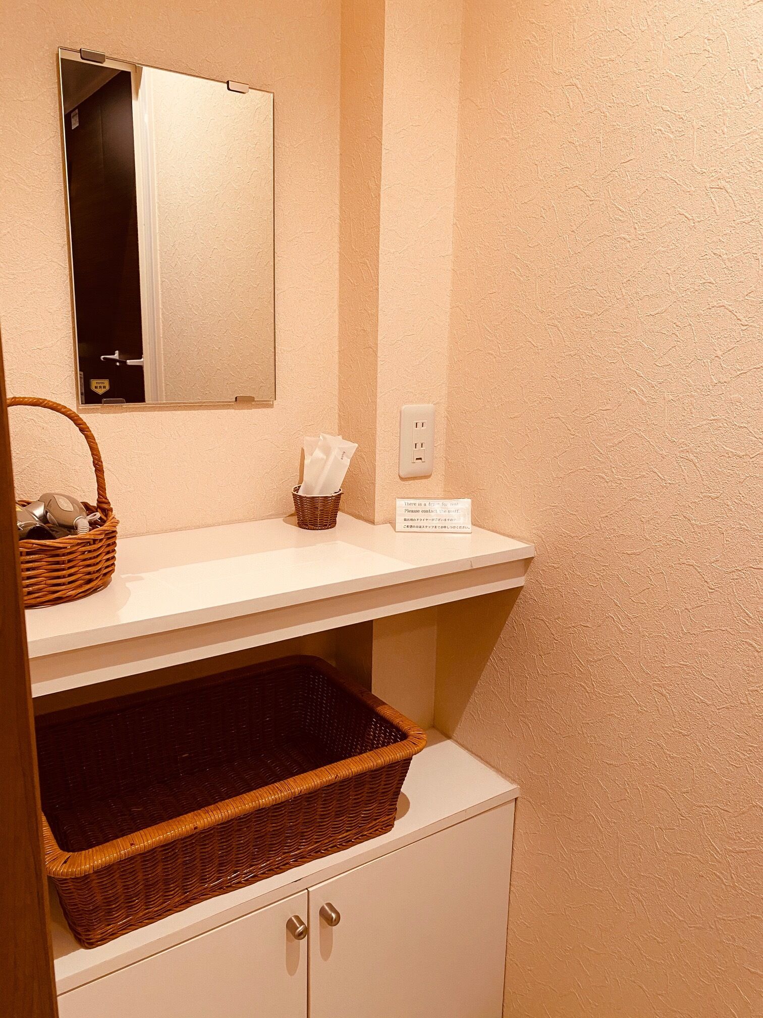 A bathroom with a mirror, a shelf with a basket and tissues, and a trash bin.