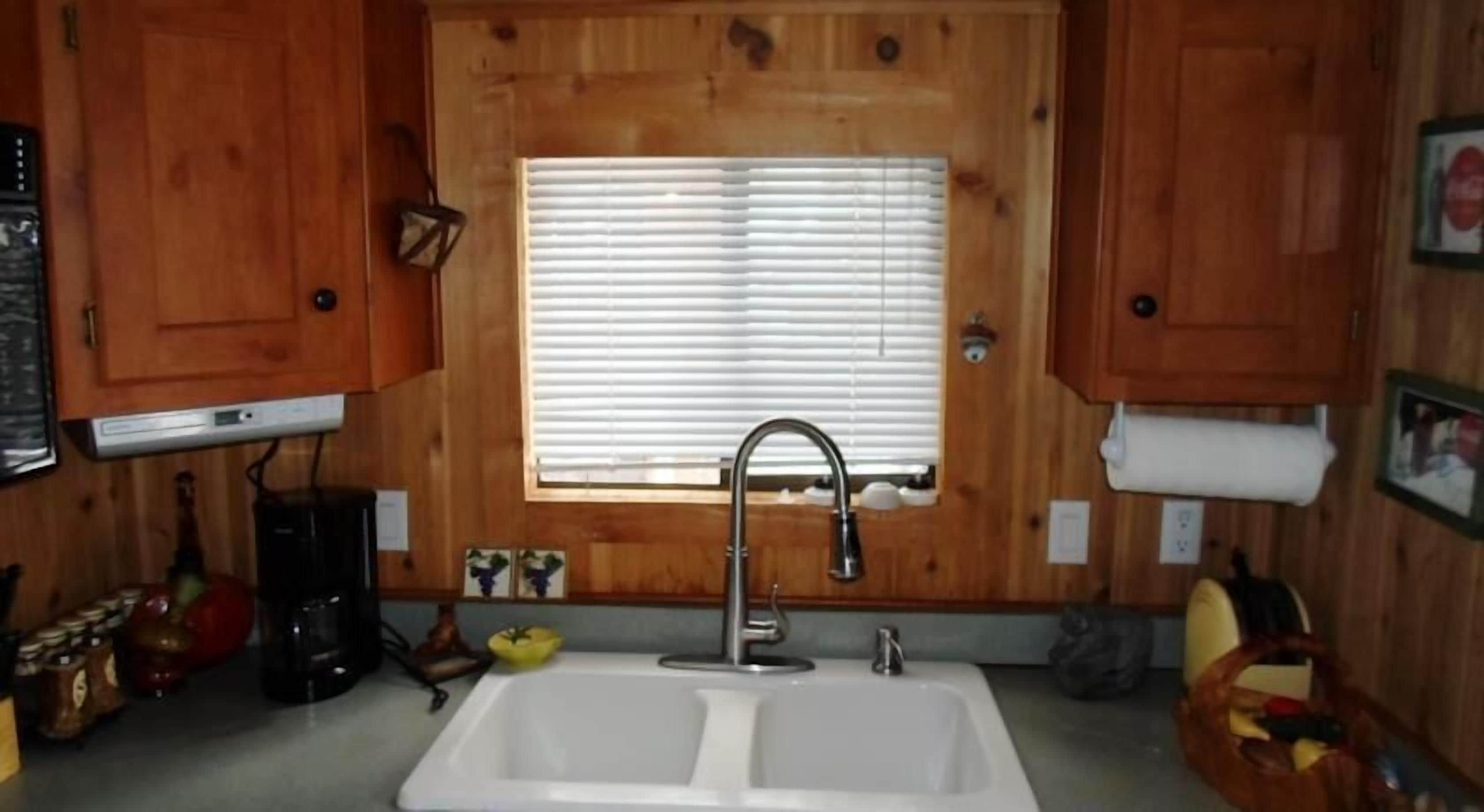 A kitchen with wooden cabinets, a double sink, a window with blinds, and a coffee maker.