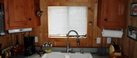 A kitchen with wooden cabinets, a double sink, a window with blinds, and a coffee maker.