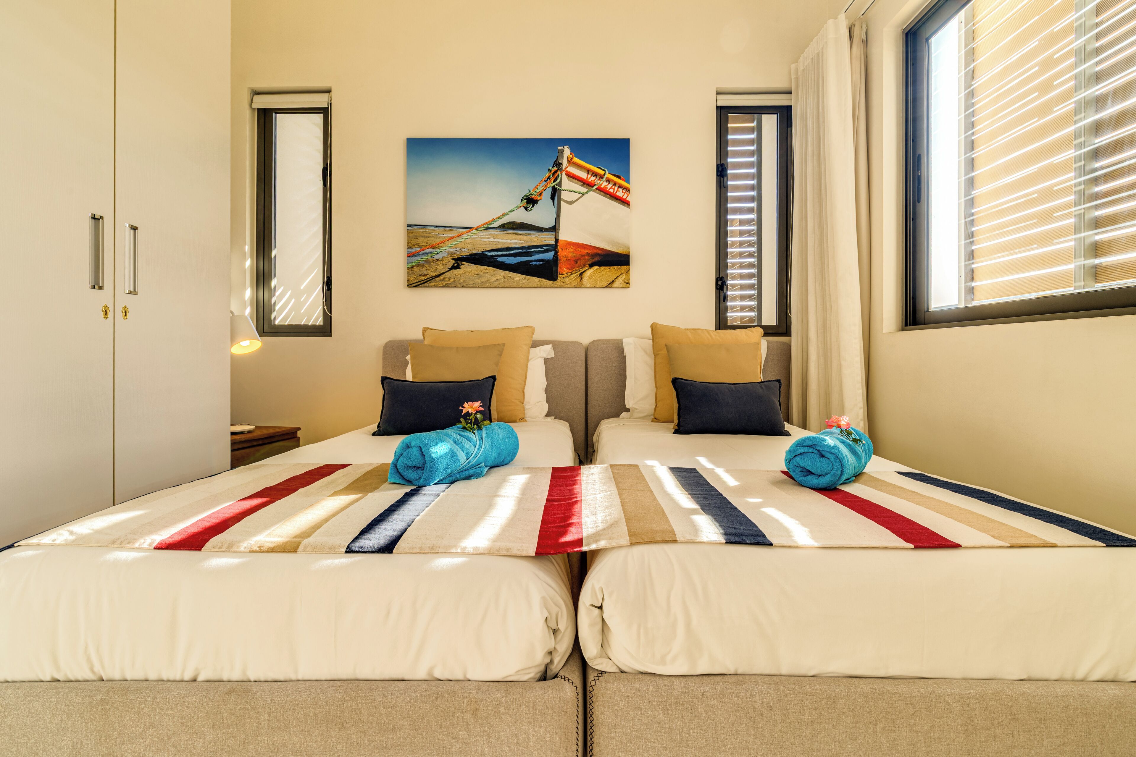 A hotel room with two beds, a painting on the wall, and a window with blinds.