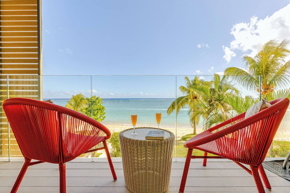 Un balcon avec deux fauteuils en osier rouge, une petite table ronde avec deux verres contenant une boisson, et une vue sur la plage et l’océan.