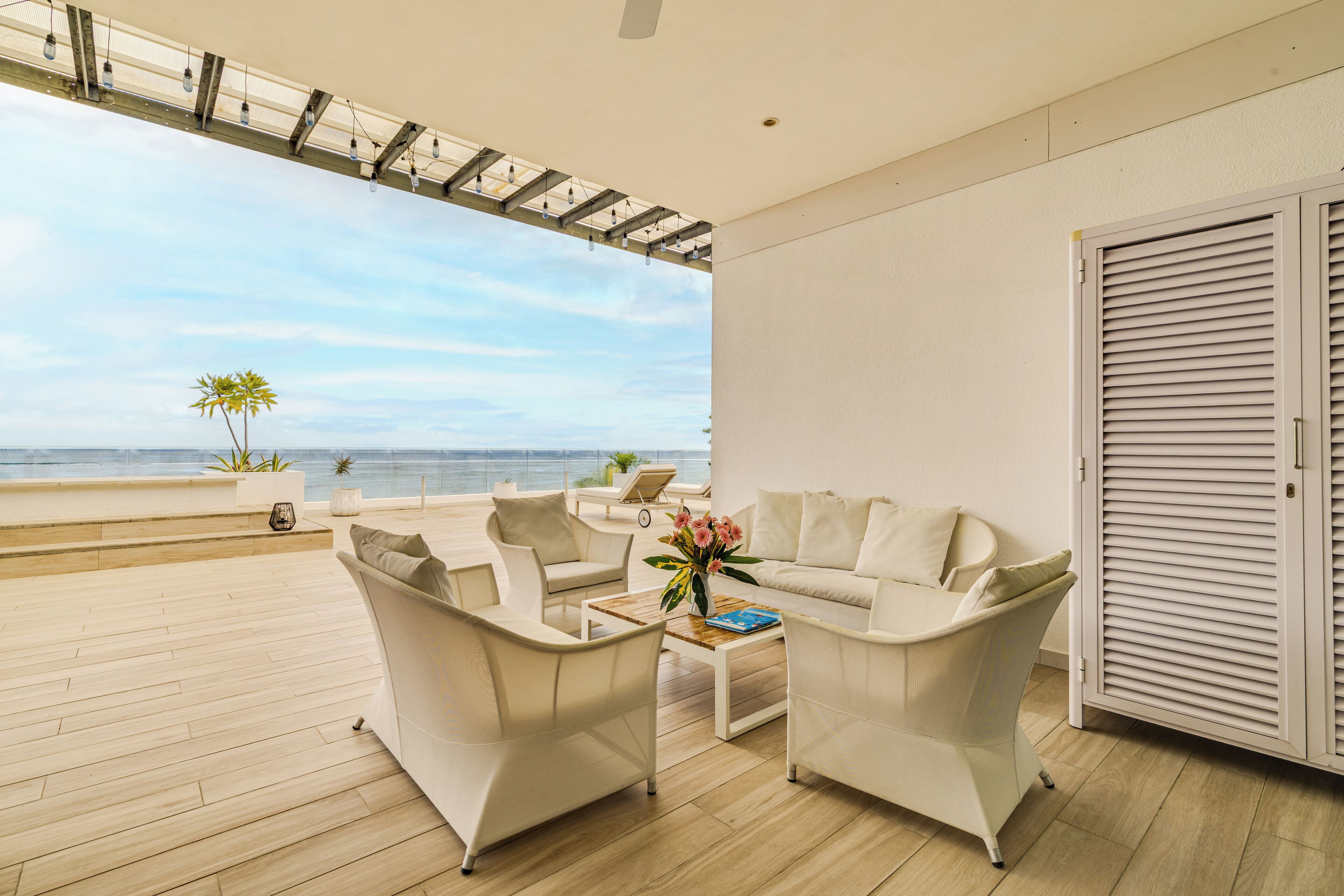 A modern living room with a wooden floor, white furniture, a coffee table, and a view of the ocean.