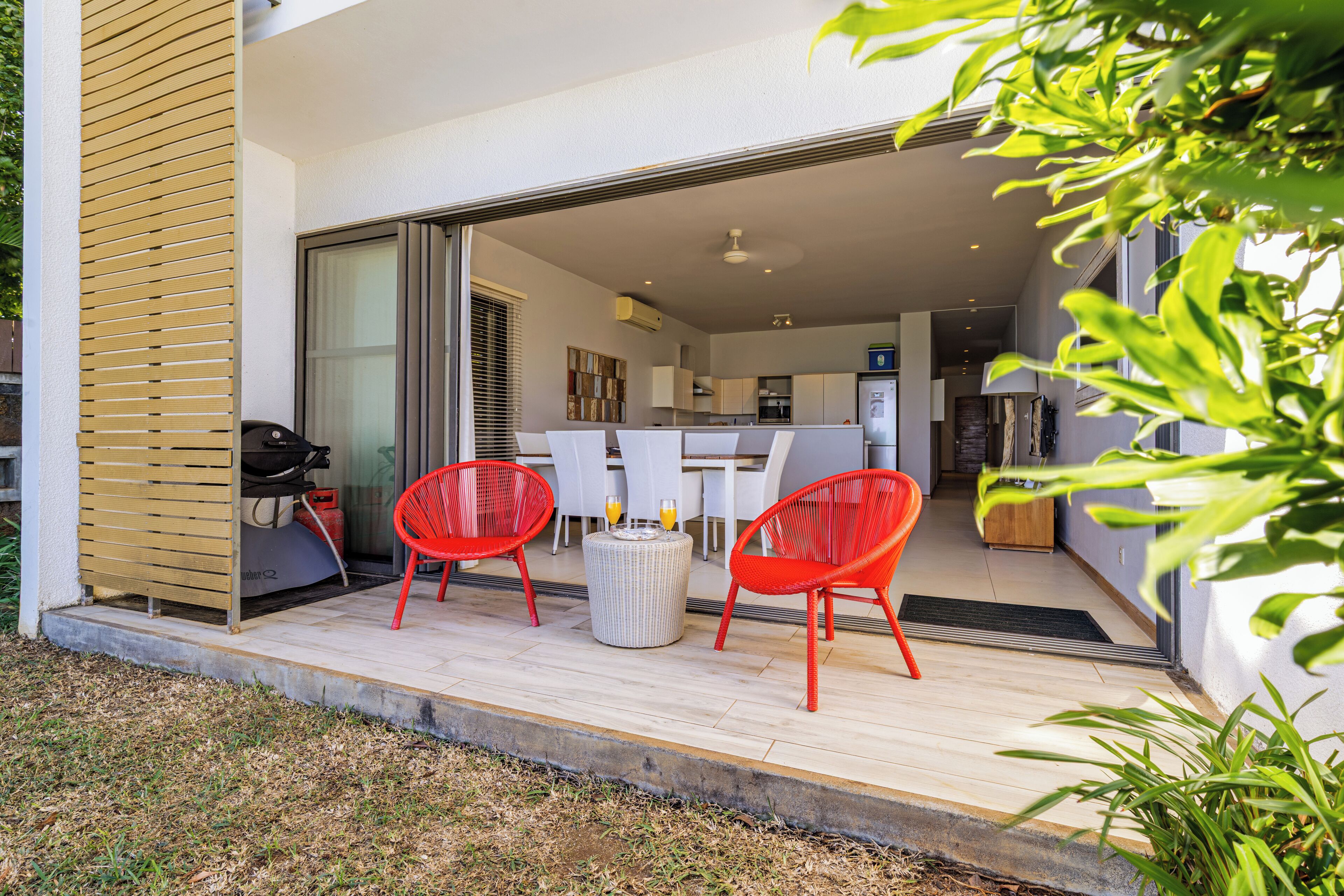 An open-plan living area with a dining table and red chairs, a kitchen in the background, and a sliding glass door leading to a balcony.