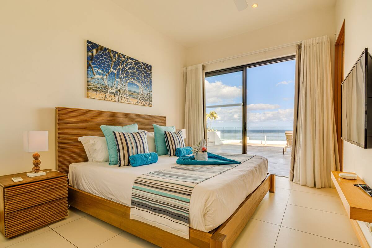 Une chambre spacieuse avec un grand lit, une tête de lit en bois, une table de chevet avec une lampe, et une vue sur la plage grâce à une porte-fenêtre coulissante.