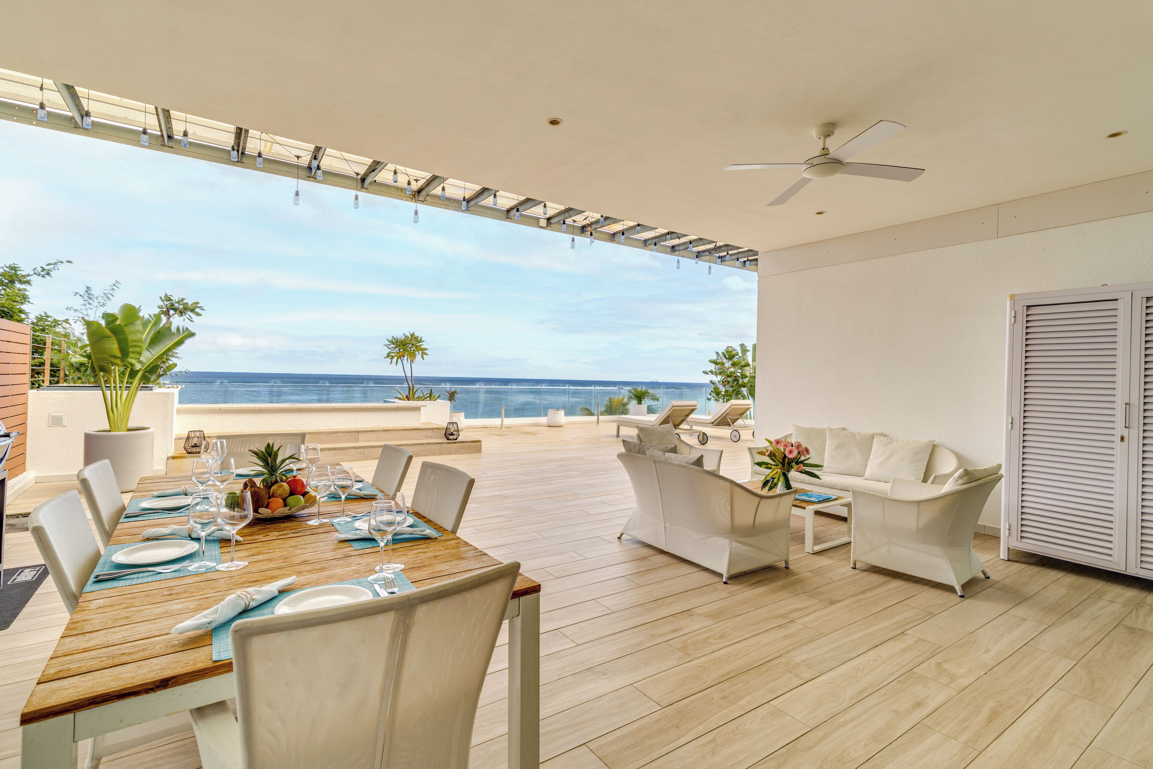 Un amplio espacio exterior con comedor, una mesa de madera para cuatro personas, sillas blancas y un sofá blanco. Se ve un ventilador de techo y un armario blanco al fondo.