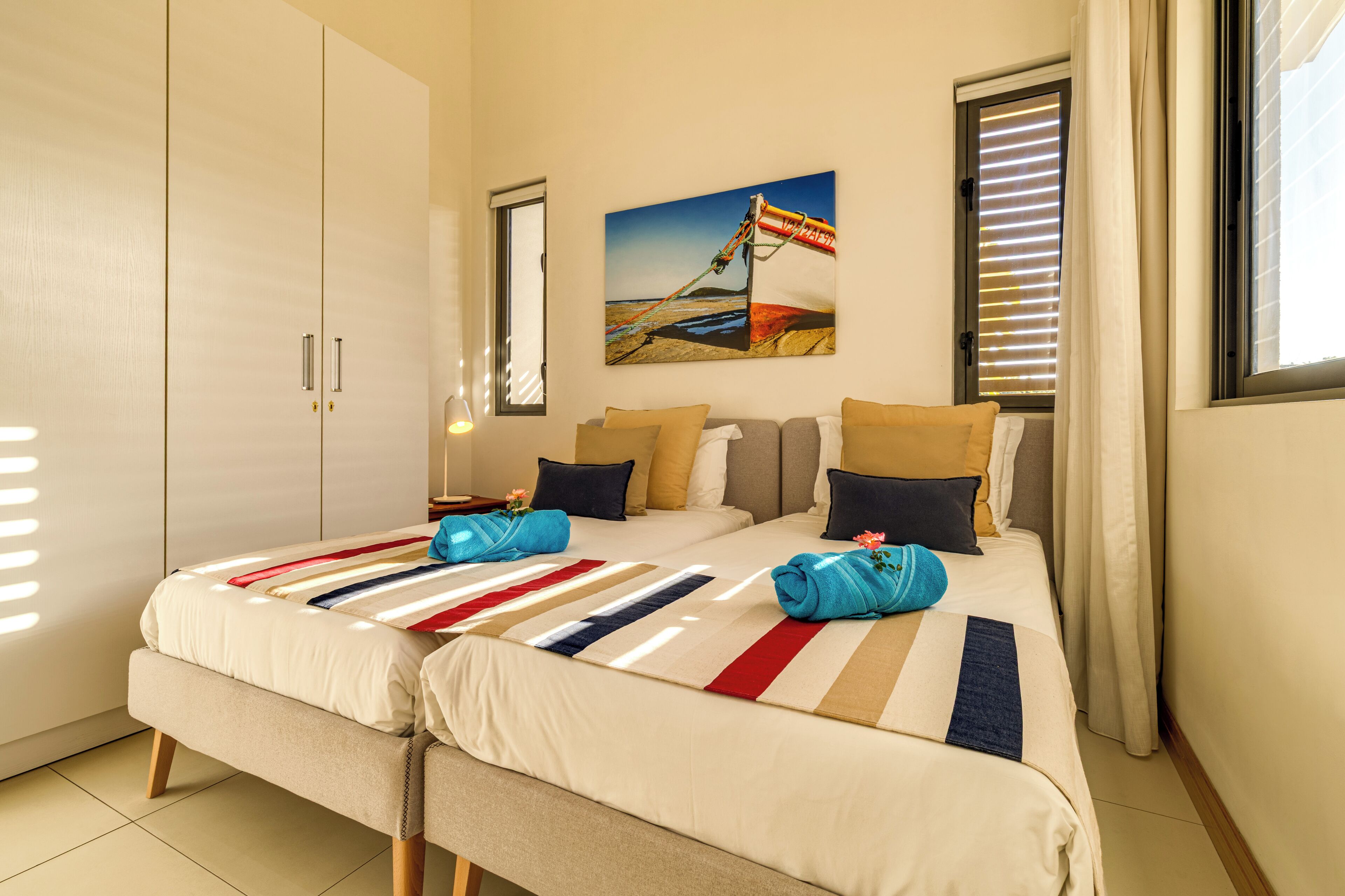A hotel room with two beds, a large window with blinds, a painting on the wall, and a wardrobe.