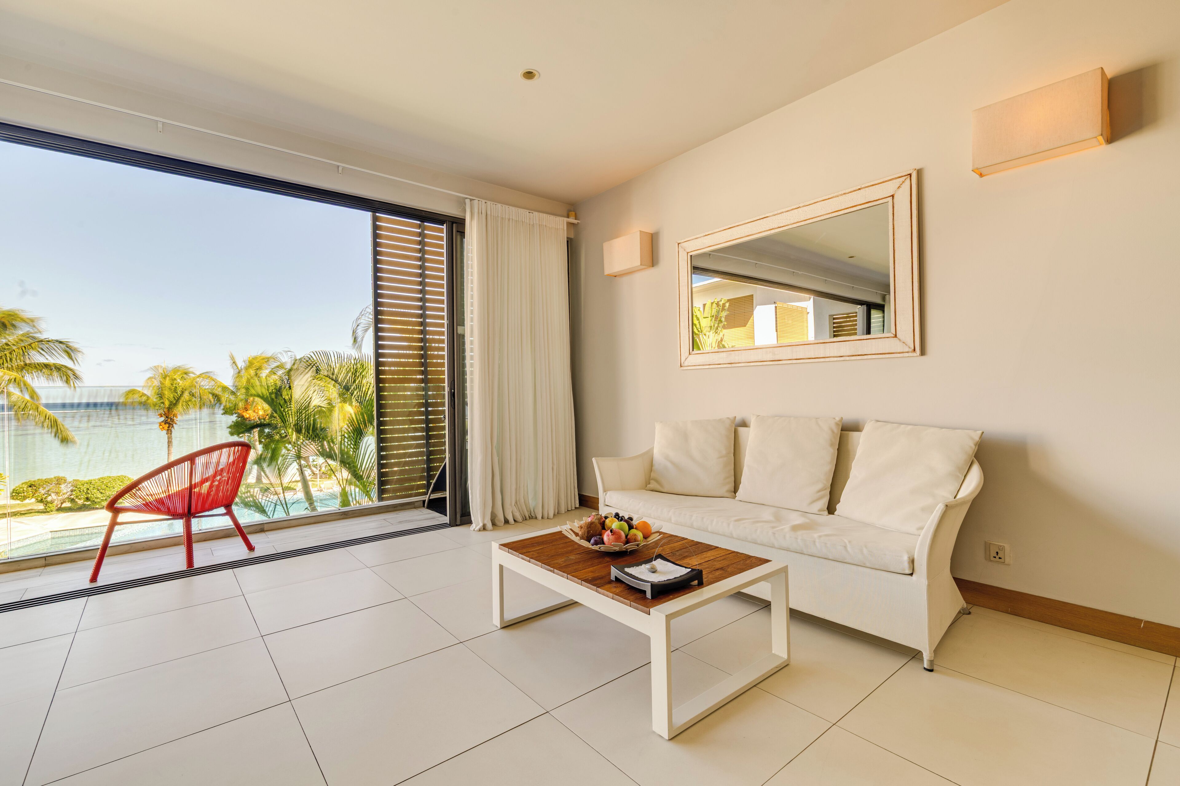 A modern living room with a white sofa, a wooden coffee table with fruit, a red chair, and a large window with a view of palm trees and the sea.