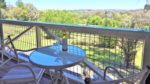 A balcony with a glass table and chairs, a bottle of wine, and a glass. The balcony overlooks a green landscape with trees and a fence. - Golden Heritage Accommodation (Beechworth)