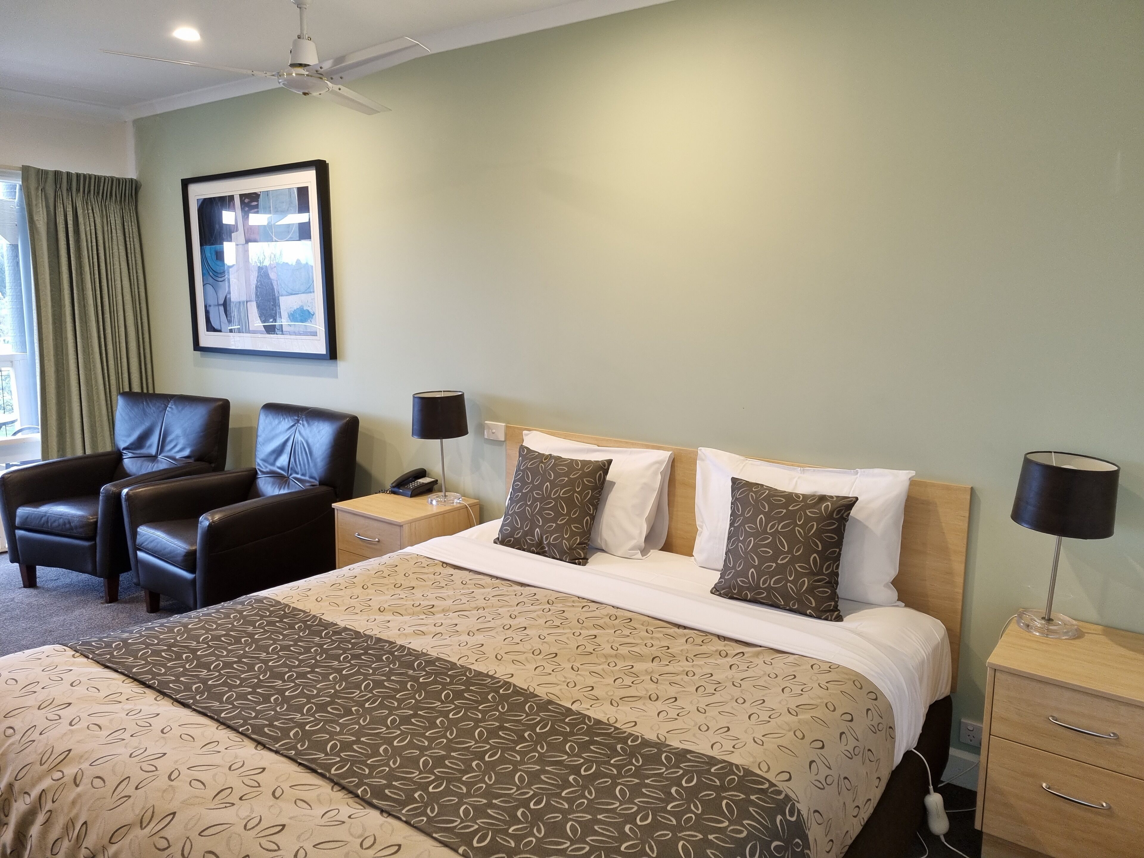 Une chambre d’hôtel avec un lit, deux chaises, une table de chevet, une lampe et un ventilateur de plafond.
