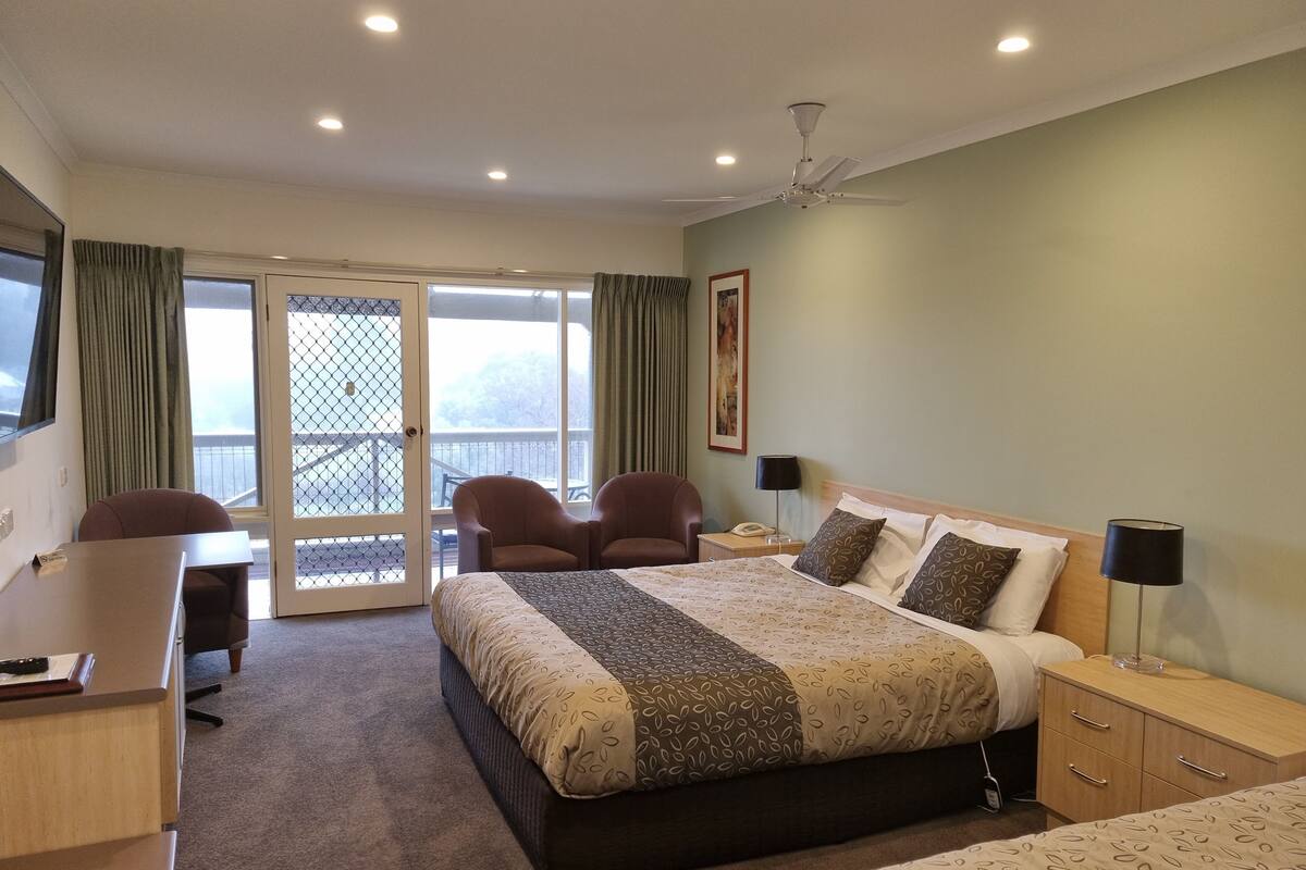 A hotel room with a bed, desk, chairs, a desk lamp, a bedside table, a telephone, and a view through the glass doors.