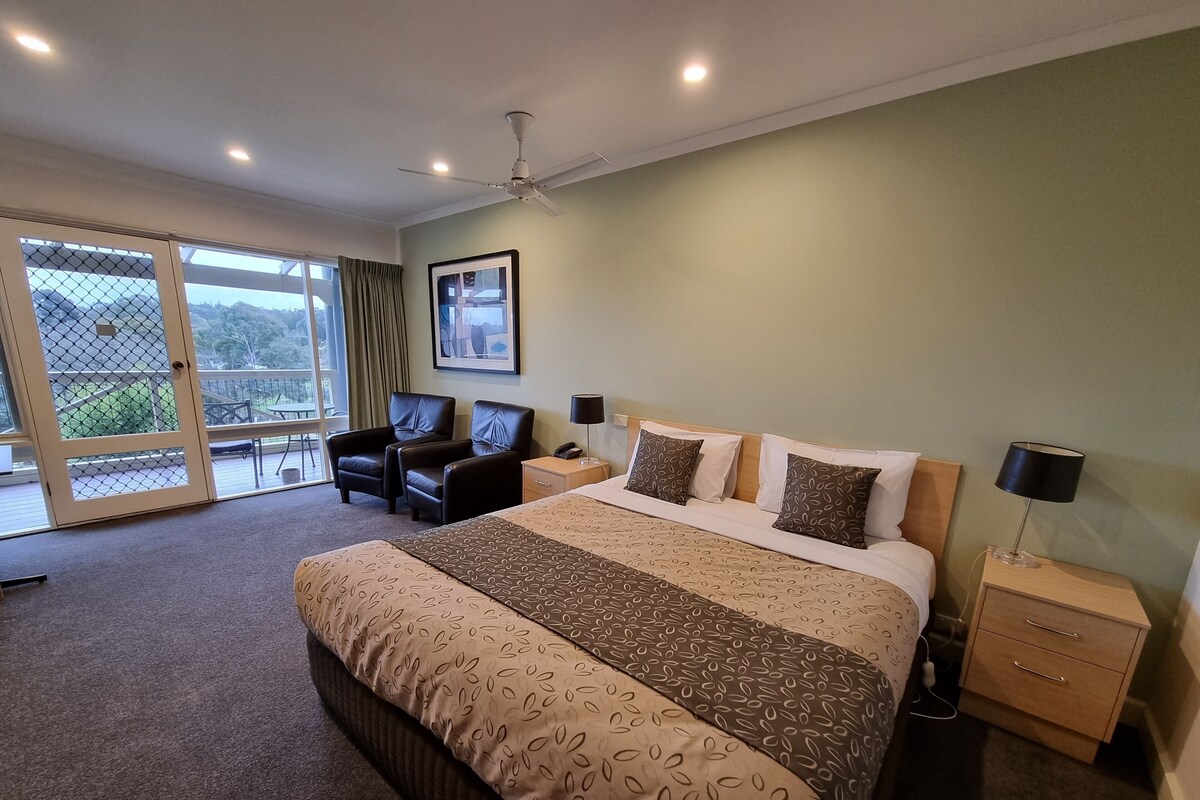 A hotel room with a large bed, two bedside tables, a ceiling fan, and a view through a glass door to an outdoor area.