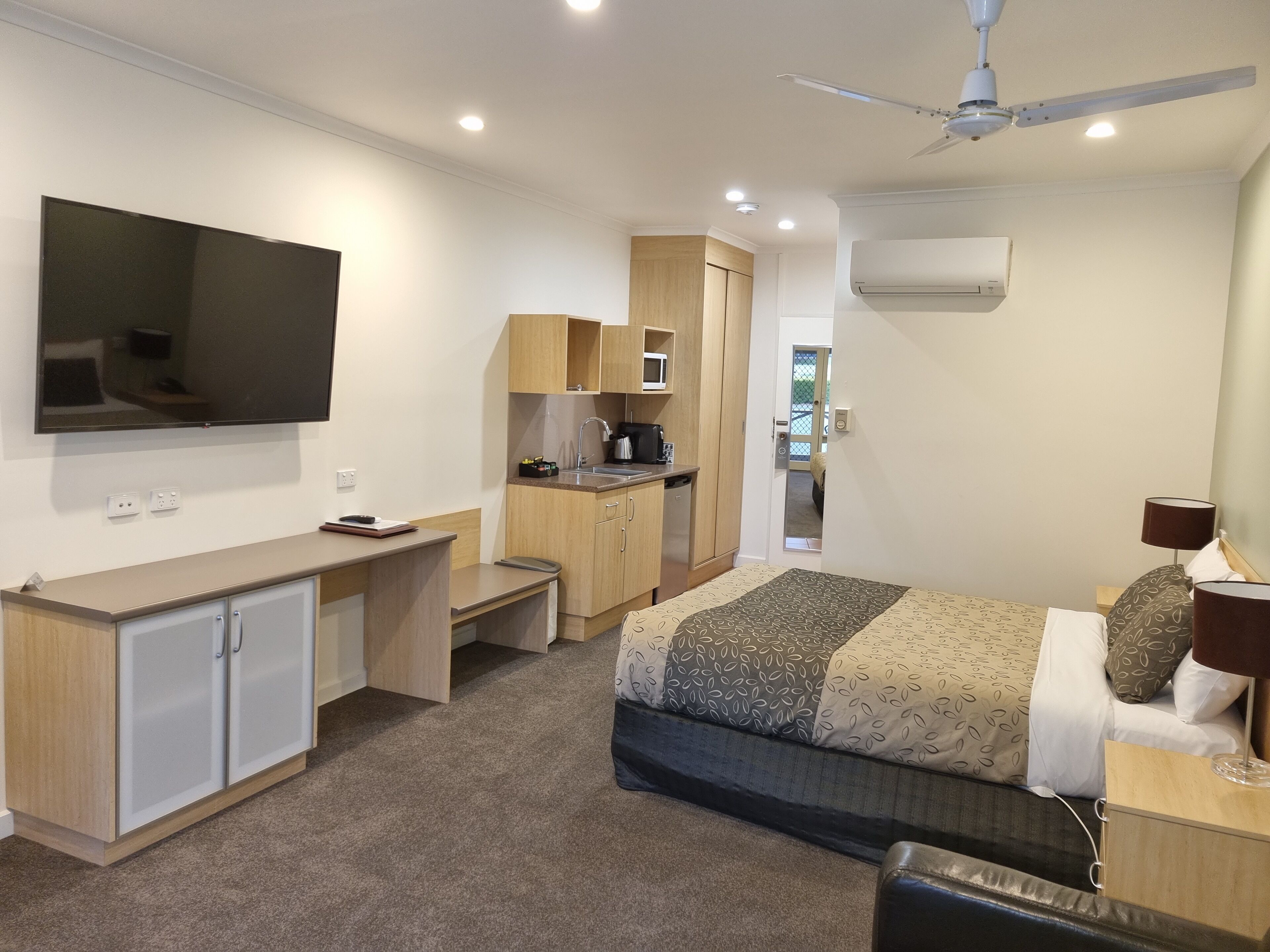 A compact hotel room with a bed, a desk, a kitchenette, and a ceiling fan.