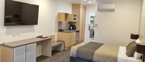 A compact hotel room with a bed, a desk, a kitchenette, and a ceiling fan.