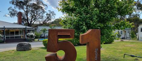 A large outdoor number '51' sign with 'Sydney Rd' inscribed on it, set against a backdrop of a residential area with houses and lush greenery.