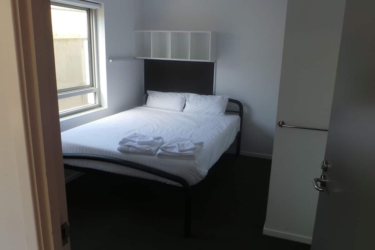 A small, neatly made bed with white linens and towels, a window with blinds, and a simple headboard.
