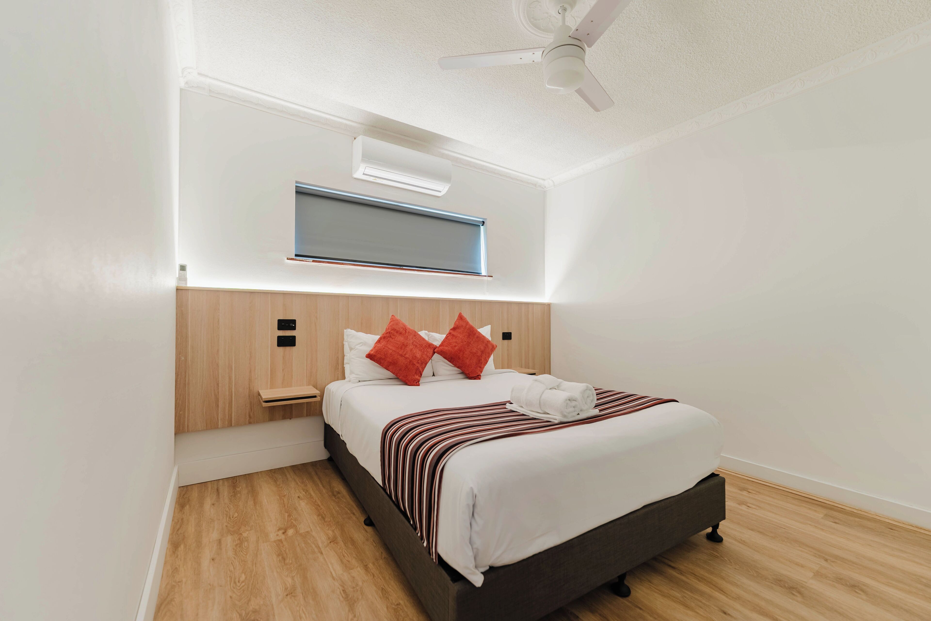 A modern hotel room with a wooden headboard, a white bed with red pillows, a striped blanket, and a ceiling fan.