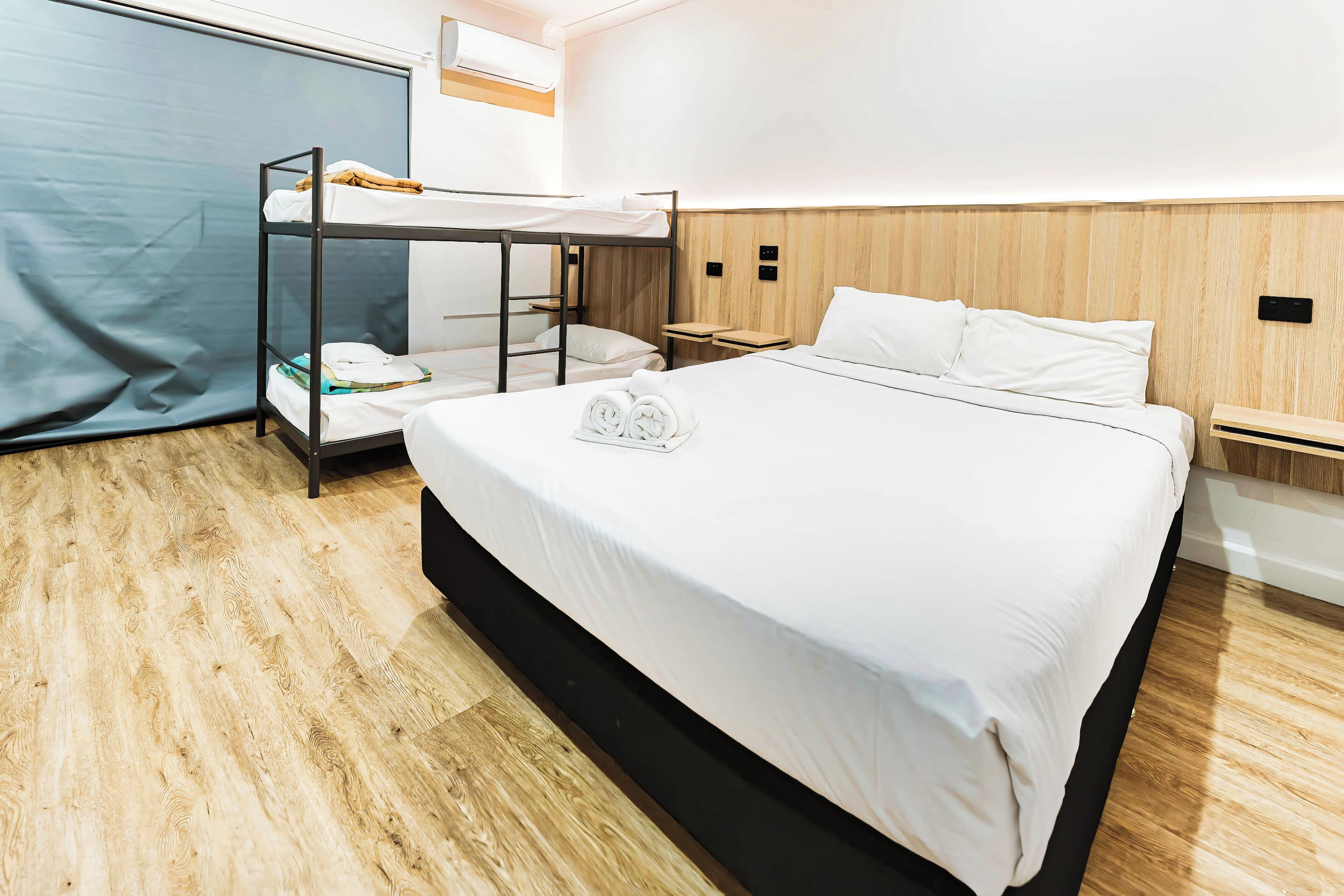 A hotel room with a bunk bed, a large bed, wooden flooring, and a wooden headboard.