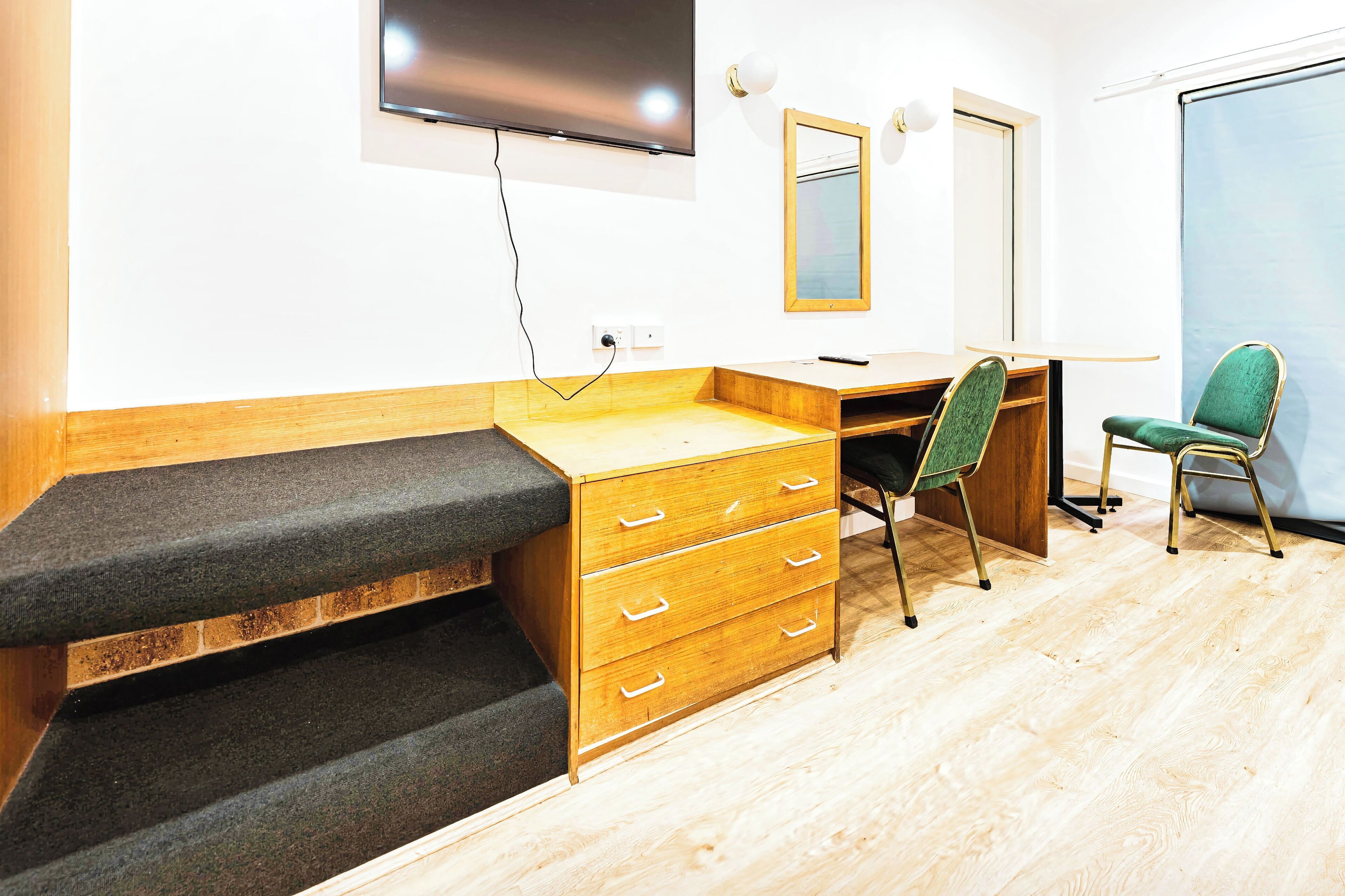 A compact room with a wooden desk, a chair, a TV mounted on the wall, and a small window.
