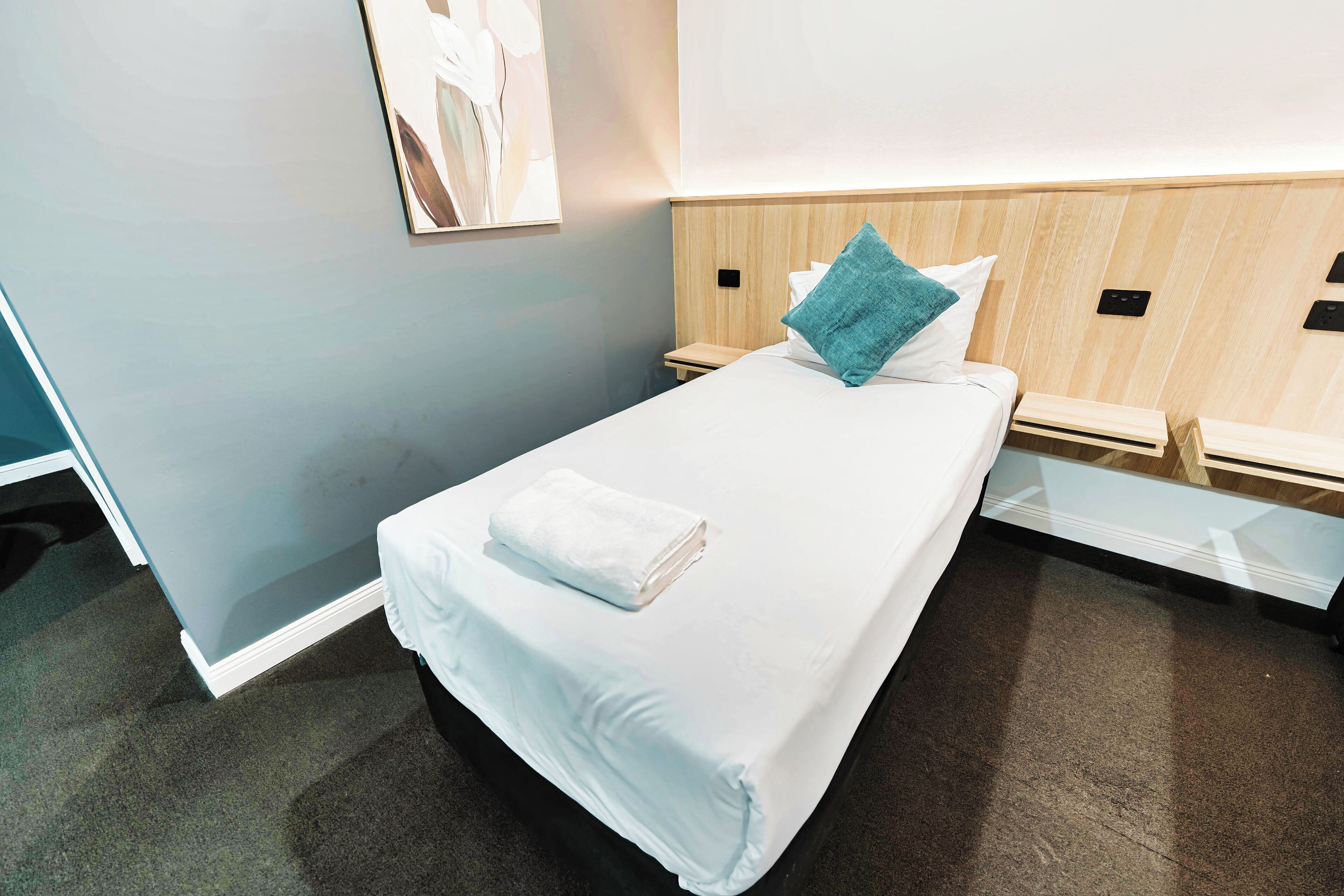 Una cama bien tendida con una colcha blanca y una toalla blanca doblada, un almohón turquesa y un cabecero de madera.