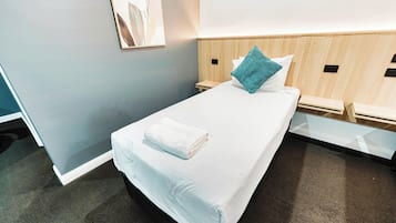 Una cama bien tendida con una colcha blanca y una toalla blanca doblada, un almohón turquesa y un cabecero de madera.