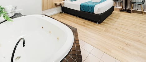 Una habitación de hotel moderna con una bañera grande, una cama bien hecha con ropa de cama color turquesa, una mesita de noche pequeña y una mesa redonda con sillas.