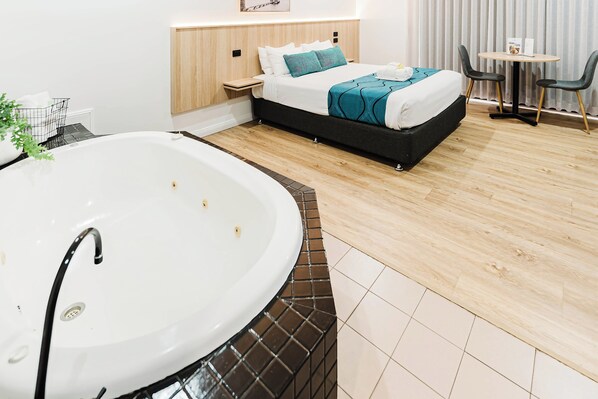 Una habitación de hotel moderna con una bañera grande, una cama bien hecha con ropa de cama color turquesa, una mesita de noche pequeña y una mesa redonda con sillas.