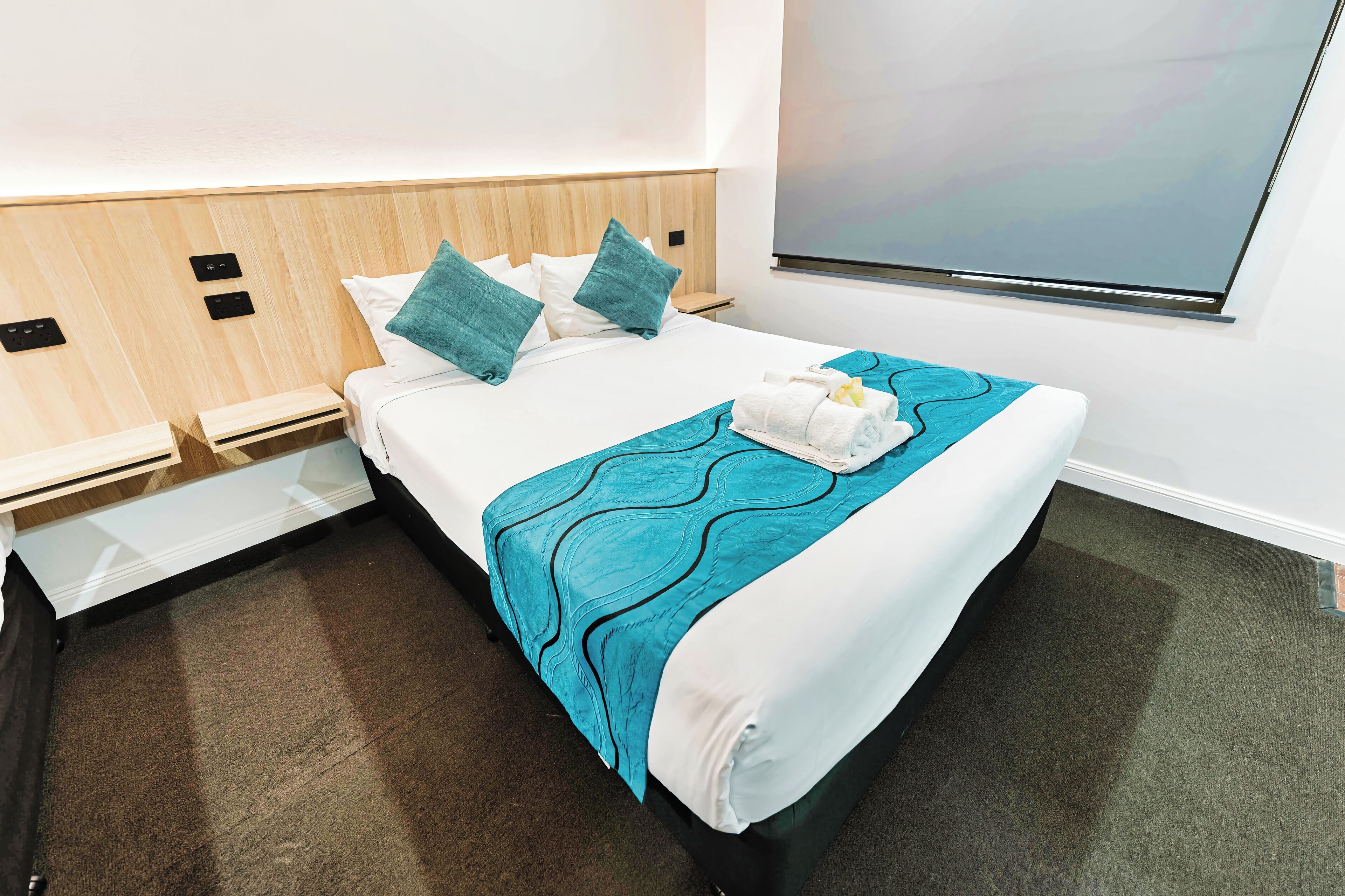 A neatly made bed with a teal blanket, white pillows, and a towel. There is a wooden headboard, a nightstand, and a wall-mounted TV.