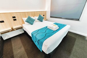 A neatly made bed with a teal blanket, white pillows, and a towel. There is a wooden headboard, a nightstand, and a wall-mounted TV.