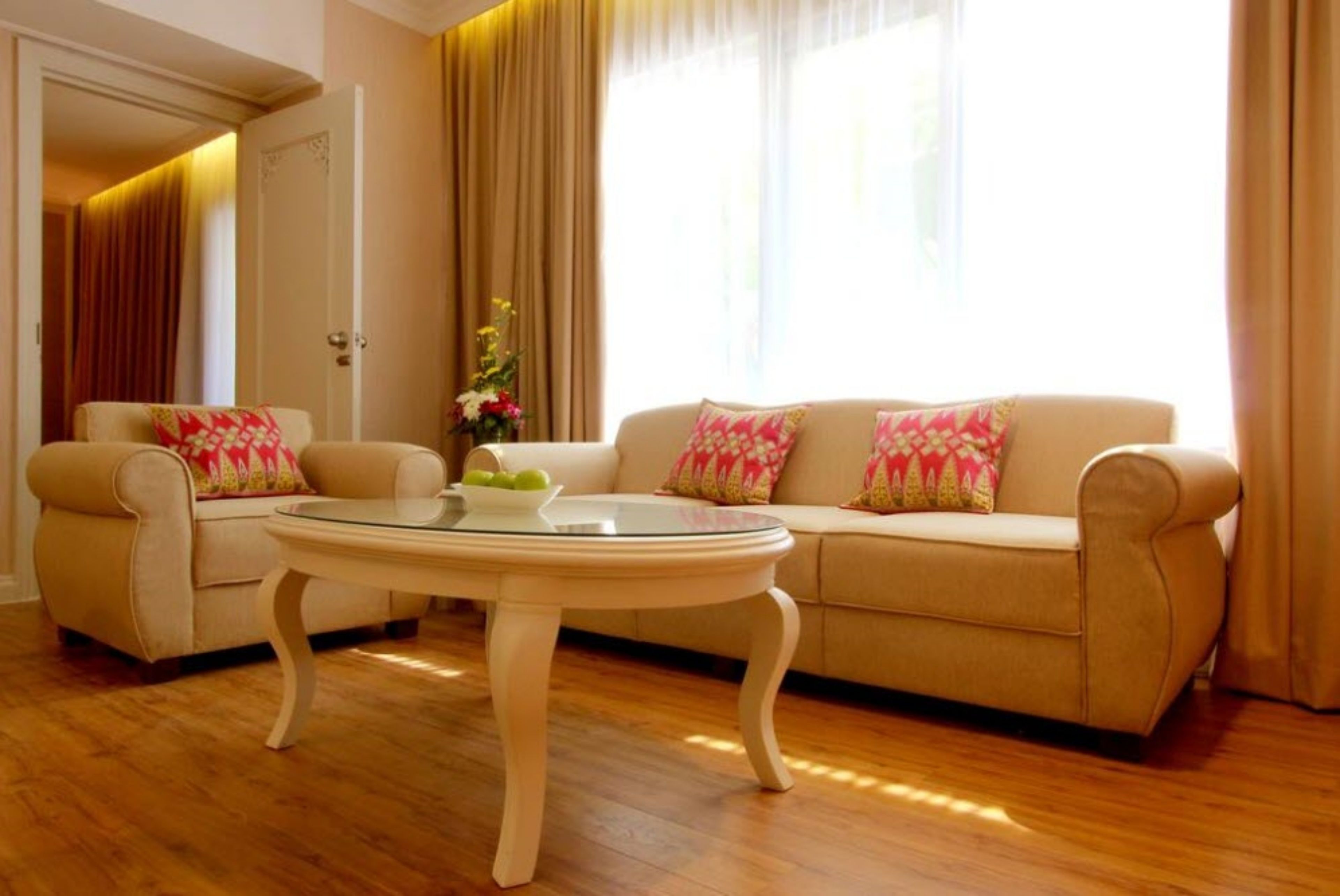 A living room with a beige sofa, two armchairs, a glass coffee table, and a door leading to another room.