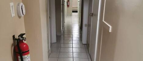 A hallway with white tiled floors, a fire extinguisher mounted on the wall, and an exit sign at the end.