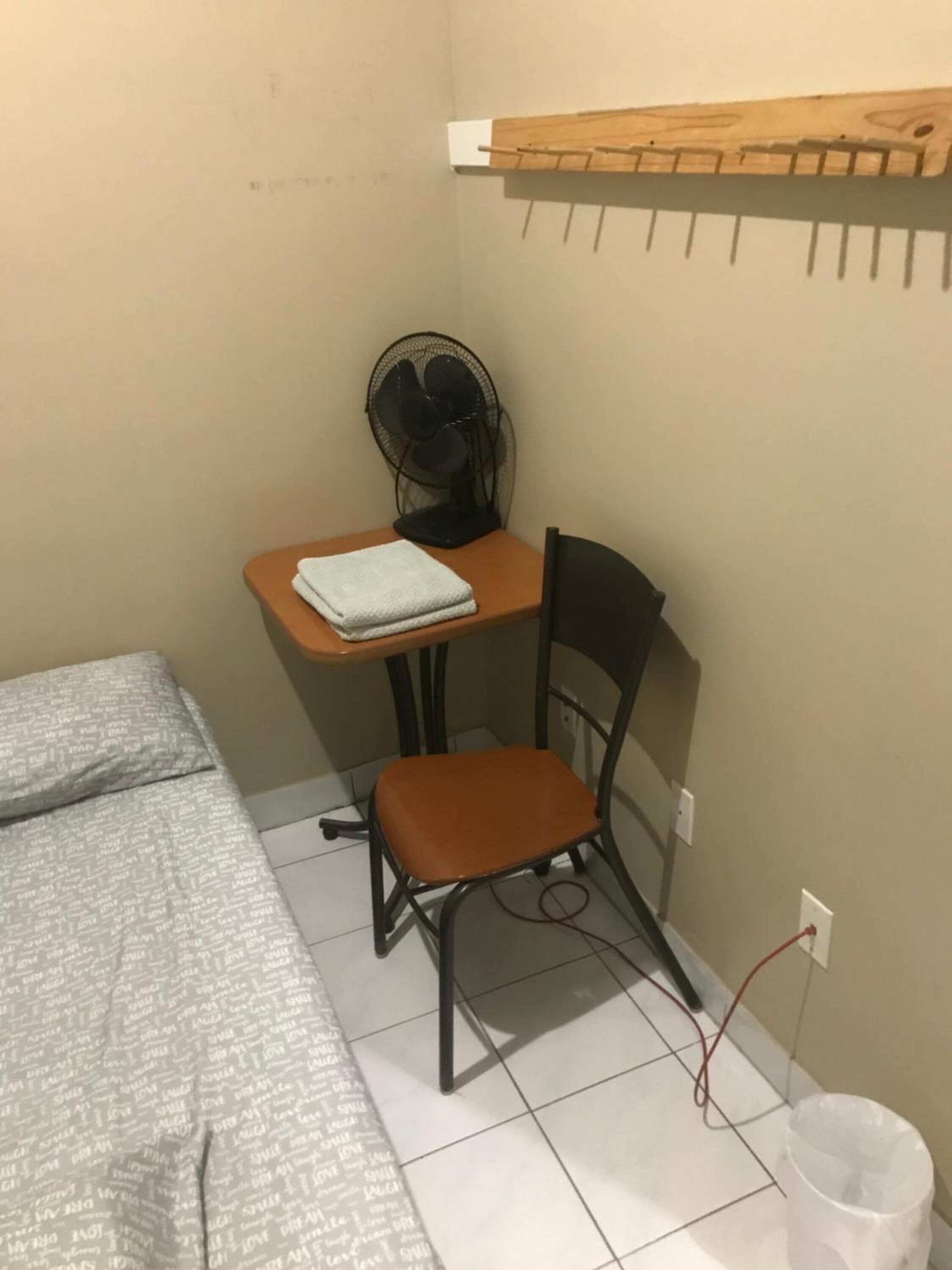 Een kleine, compacte kamer met een enkel bed, een bureau met stoel, een ventilator en een kleine prullenbak.