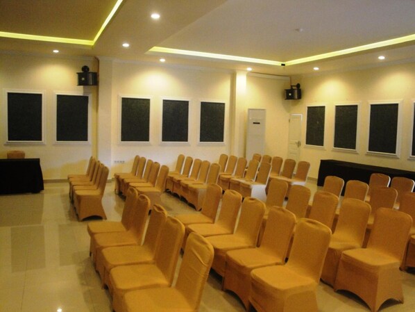 Un salón de conferencias con filas de sillas y mesas, ventanas amplias y un techo plano con iluminación empotrada.