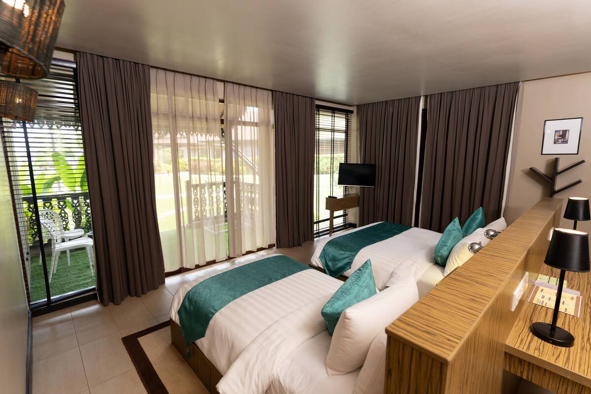 A hotel room with two beds, a wooden bedside table, a TV, and a balcony with a view of greenery.