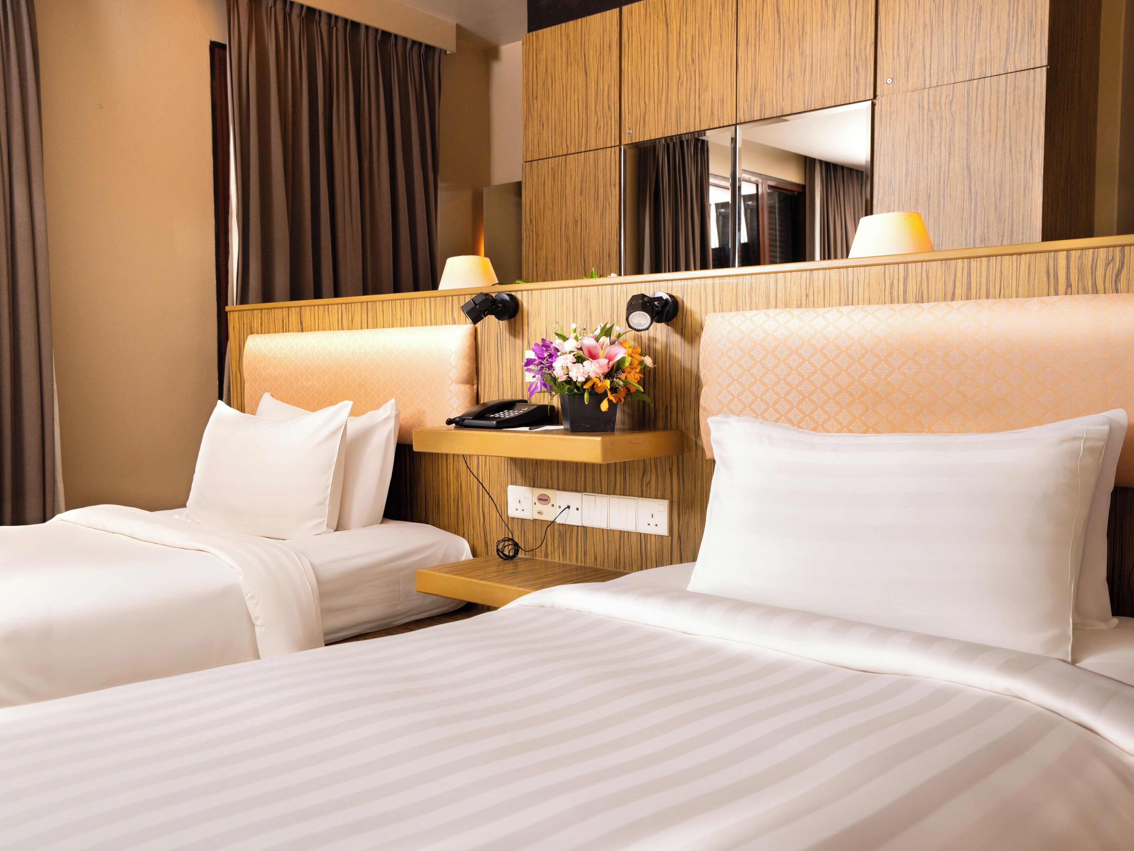A hotel room with two beds, a wooden headboard, a bedside table with a lamp, a phone, and a vase of flowers.