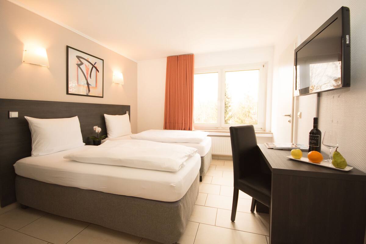 A hotel room with two beds, a desk with a bottle and fruit, and a window with curtains.