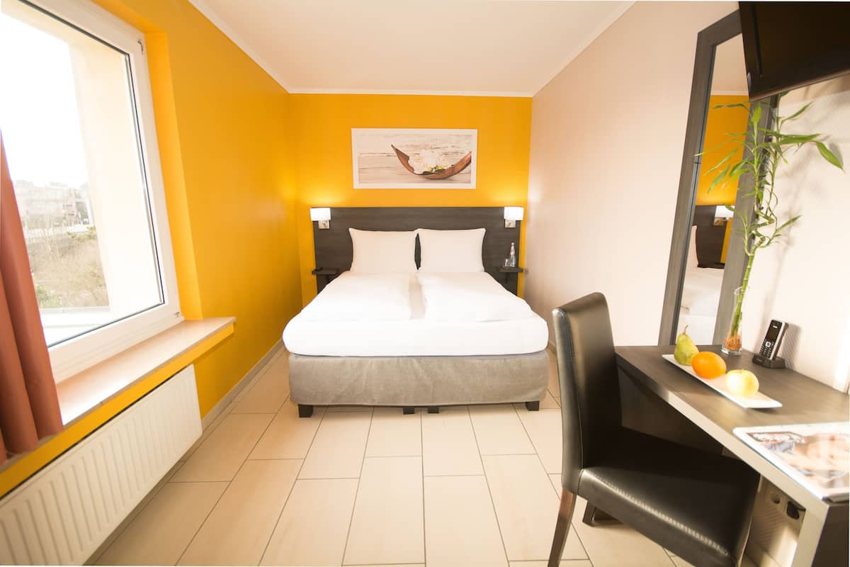 A hotel room with a bed, a desk with a chair, a window, and a mirror.