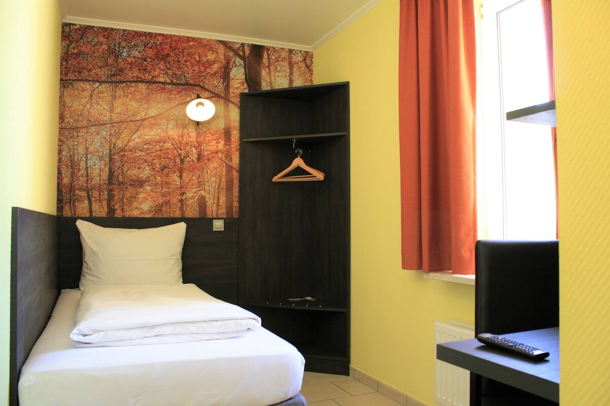 A hotel room with a bed, a desk, a hanging coat rack, and a window with curtains.